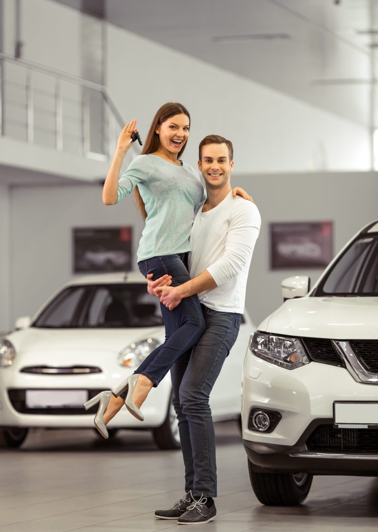 Beautiful young couple is smiling, looking at camera and showing keys while buying a car in a motor show. Man is holding woman in his arms