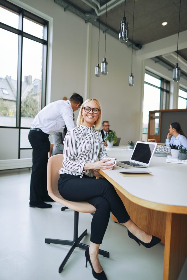 Young businesswoman smiling and drinking a coffee while sitting at a boardroom table with colleagues working in the background